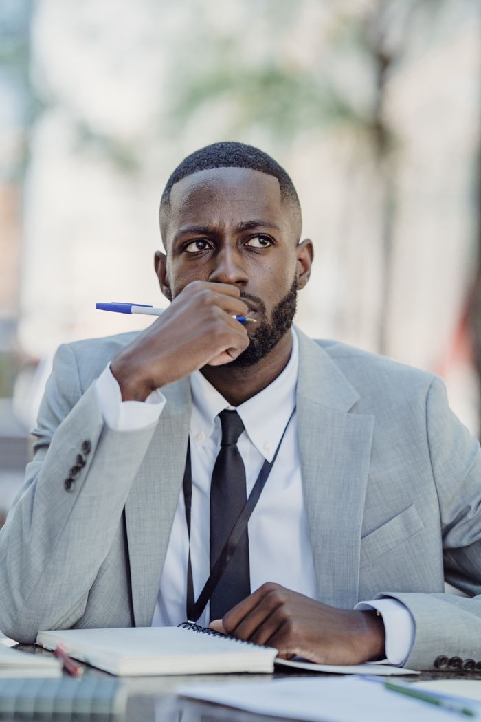 A businessman in a grey suit sits outdoors pondering with a pen and notebook.
