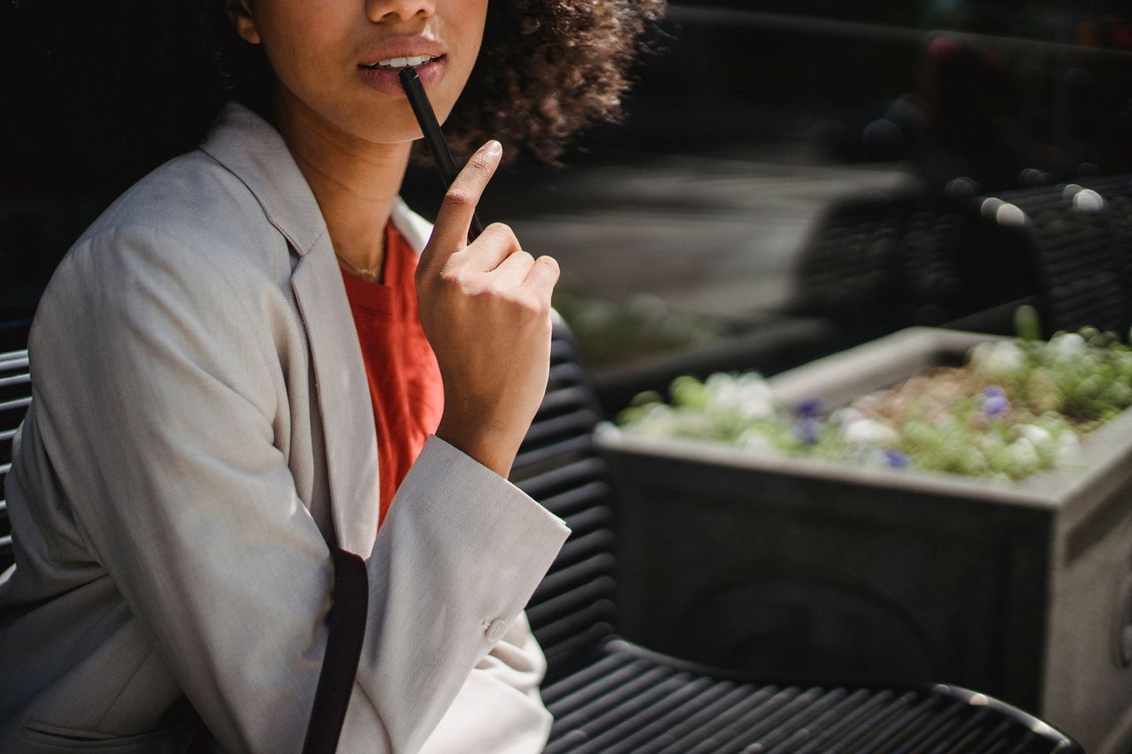 Stylish woman in a blazer holds a pen while sitting outdoors, deep in thought.