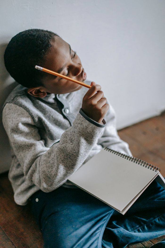 High angle of African American boy resting on floor with blank notebook and scratching head with pencil