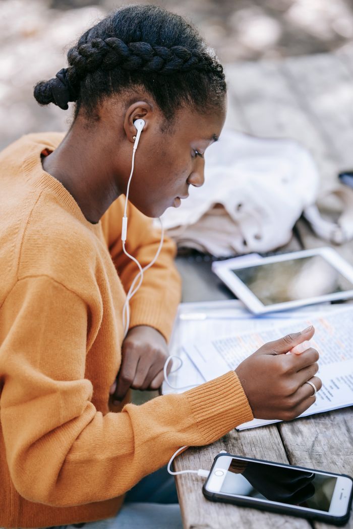 Side view of serious young African American female in earphones learning information while studying at table with mobile phone