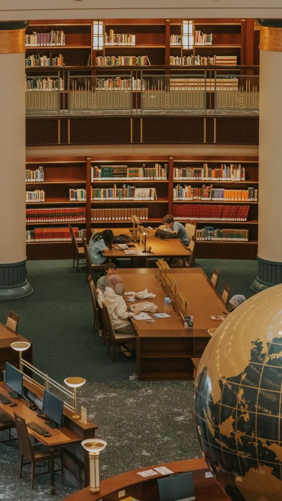 Students studying at tables in a university library surrounded by bookshelves and a globe.