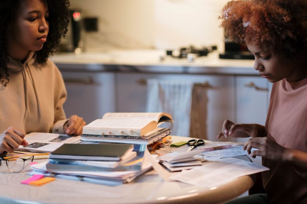 Two young women study together at a table filled with books and papers, focused on learning.