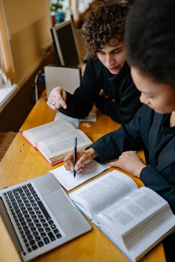 Two students studying together with books and a laptop, taking notes in a cozy indoor setting.
